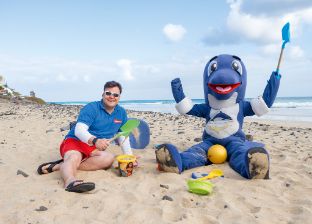 Mann sitzt mit Flosse am Strand und hält Sandspielzeug in der Hand 