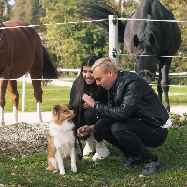Zwei Personen mit Hund und im Hintergrund sind Pferde 
