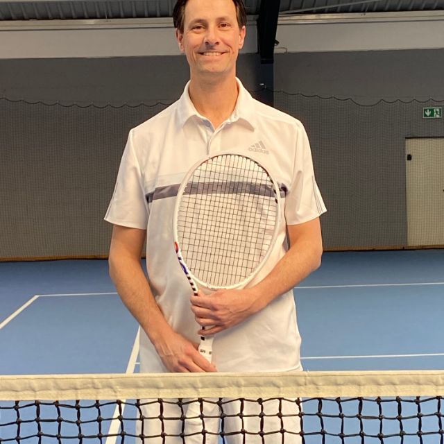 Ein Mann lächelt in die Kamera und hält einen Tennisschläger in der Hand